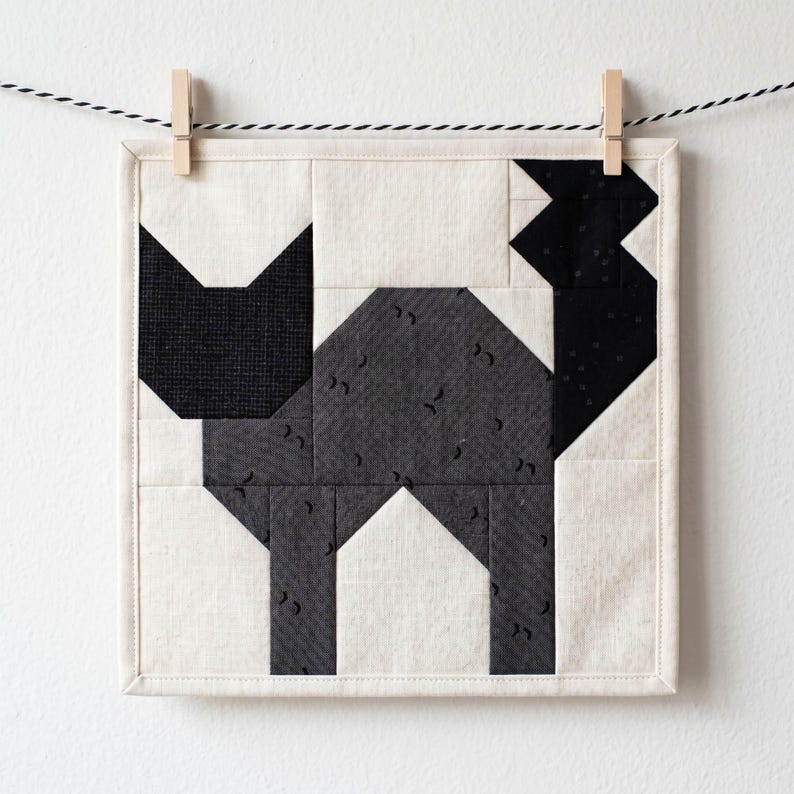 May include: A square, quilted wall hanging featuring a geometric cat design. The cat is composed of black and gray fabric pieces on a cream background. The artwork is suspended by a clothes pin on a black and white string.