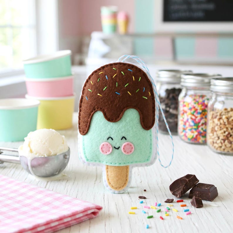May include: A felt ice cream popsicle with a mint green base, brown chocolate topping, and colorful sprinkles. The popsicle has a smiling face and a wooden stick. Other items in the image include ice cream, cups, and jars of toppings.