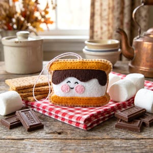 May include: A felt s'more ornament with a smiling face, graham cracker, chocolate, and marshmallows on a red and white checkered cloth. The s'more has a brown chocolate layer, a white marshmallow layer, and a yellow graham cracker layer.