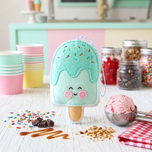 May include: A felt ice cream popsicle ornament with a smiling face, mint green top, and colorful sprinkles. The ornament is on a white surface with ice cream, toppings, and paper cups.