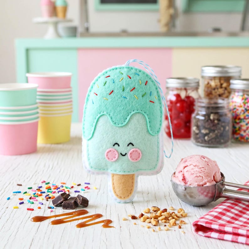 May include: A felt ice cream popsicle ornament with a smiling face, mint green top, and colorful sprinkles. The ornament is on a white surface with ice cream, toppings, and paper cups.