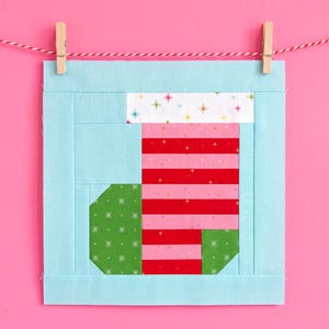 Peut inclure: Un motif de chaussette de Noël matelassée carrée, avec des rayures rouges et roses, des motifs géométriques verts et un revers blanc avec des étoiles colorées. Le motif est encadré en bleu clair et suspendu avec une pince à linge sur une ficelle rouge et blanche sur fond rose.