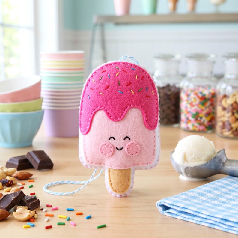 May include: A handmade felt popsicle with a smiling face. The pink popsicle has a darker pink top with colorful sprinkles and a light pink base. A light blue string is attached to the top.