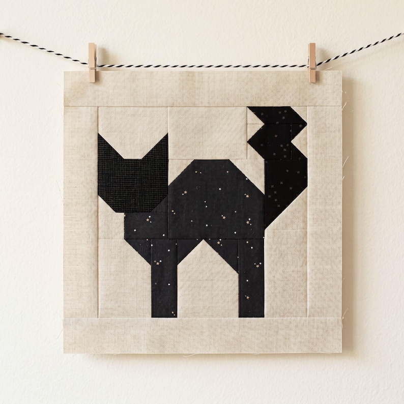 May include: A square, fabric quilt block featuring a geometric black cat design. The cat is set against a light beige background and is suspended by a clothesline with wooden clothespins. The cat's body has a speckled pattern.