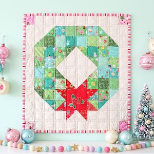 May include: A square quilted wall hanging featuring a central wreath design made of green and multicolored holiday-themed fabric squares. A large red star with a poinsettia print is at the bottom. The border is cream with red and white stripes.
