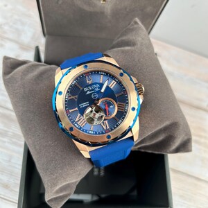 Pu&ograve; includere: Un orologio automatico Bulova Marine Star con quadrante blu e cassa color oro. L'orologio presenta indici delle ore con numeri romani, un movimento a vista e un cinturino in gomma blu. L'orologio &egrave; presentato in una scatola nera con un cuscino grigio.