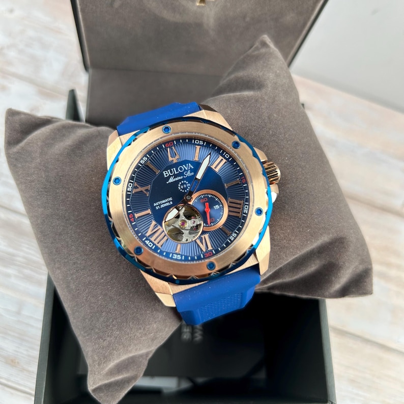 Pu&ograve; includere: Un orologio automatico Bulova Marine Star con quadrante blu e cassa color oro. L'orologio presenta indici delle ore con numeri romani, un movimento a vista e un cinturino in gomma blu. L'orologio &egrave; presentato in una scatola nera con un cuscino grigio.
