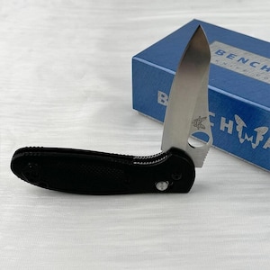 May include: A black folding knife with a silver blade, partially open, featuring a round hole near the base. The knife is set against a blue box with white text that reads "BENCHMADE KNIFE CO."