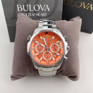 May include: A silver-tone stainless steel Bulova Marine Star chronograph watch featuring a vibrant orange dial, three sub-dials, luminous hands, and a date window. The watch has a metallic link bracelet and is displayed on a brown cushion.