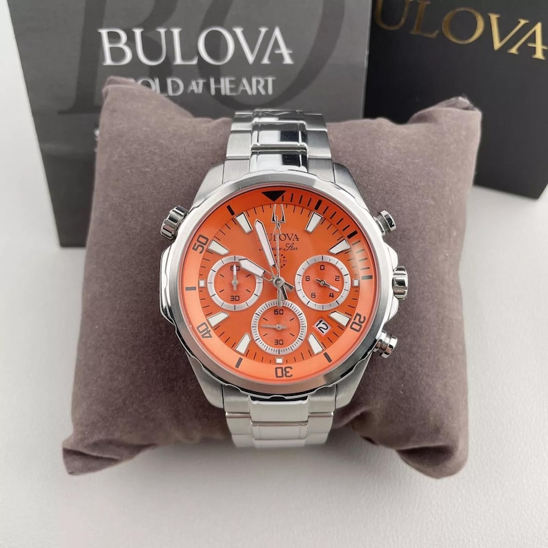May include: A silver-tone stainless steel Bulova Marine Star chronograph watch featuring a vibrant orange dial, three sub-dials, luminous hands, and a date window. The watch has a metallic link bracelet and is displayed on a brown cushion.