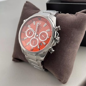 May include: A stainless steel Bulova chronograph watch with a vibrant orange dial and silver accents. The watch features multiple sub-dials and a matching stainless steel band. The watch is displayed on a brown pillow.