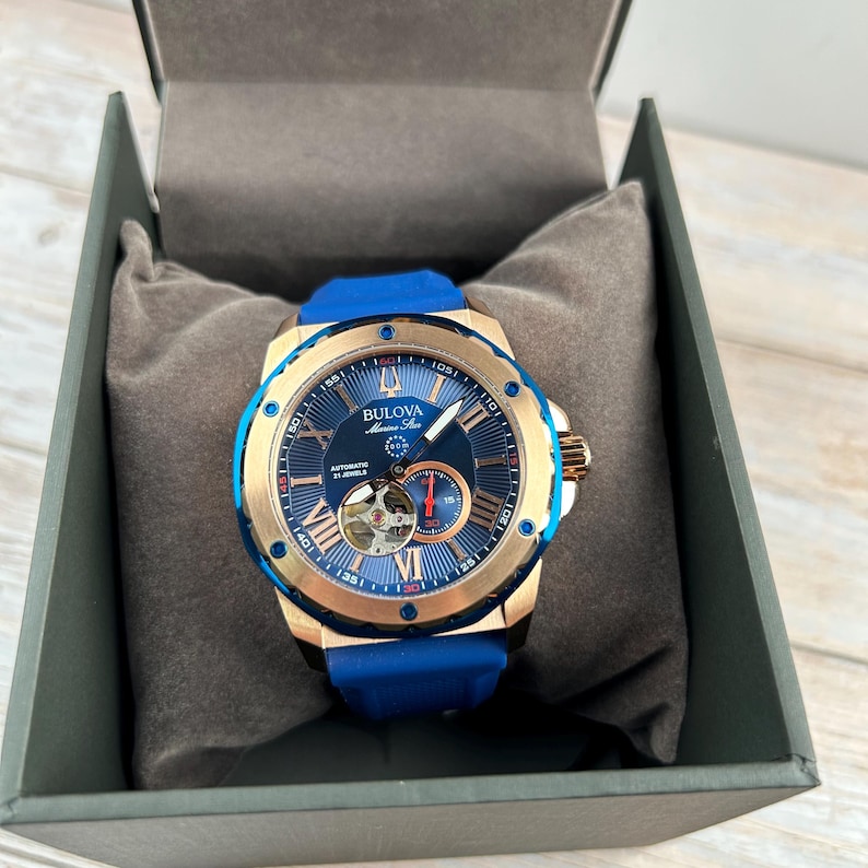 Pu&ograve; includere: Un orologio automatico Bulova Marine Star con un design blu e oro. L'orologio presenta un quadrante blu con numeri romani, un cinturino in silicone blu ed &egrave; presentato in una scatola grigia.