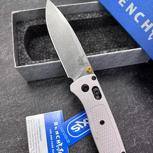 Könnte beinhalten: Ein silbernes Klappmesser mit grauem Griff und Benchmade-Logo. Das Messer befindet sich in einer grauen Box mit blauem Deckel mit der Aufschrift "Benchmade". Ein blaues Etikett mit dem Text "SIX" und "Benchmade" ist ebenfalls sichtbar.