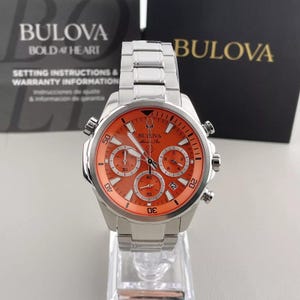 May include: A Bulova men's watch with a stainless steel band and an orange face. The watch features multiple subdials and the Bulova logo. The watch is displayed on a clear stand, with setting instructions and warranty information in the background.