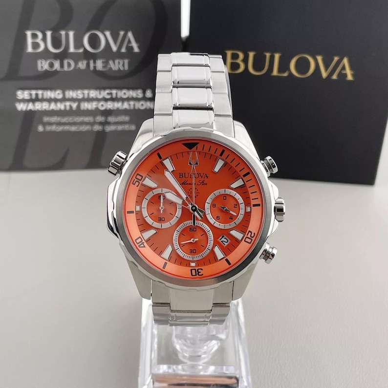 May include: A Bulova men's watch with a stainless steel band and an orange face. The watch features multiple subdials and the Bulova logo. The watch is displayed on a clear stand, with setting instructions and warranty information in the background.