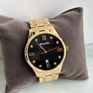 May include: A gold-tone Bulova wristwatch featuring a round black dial with diamond hour markers, Roman numerals at the twelve, three, six, and nine o'clock positions, a date window at the six o'clock position, and a matching gold-tone metal link band.