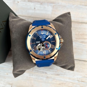 Pu&ograve; includere: Un orologio automatico Bulova Marine Star con quadrante blu, cassa color oro e cinturino in gomma blu. L'orologio presenta numeri romani e un movimento a vista. L'orologio &egrave; esposto su un cuscino grigio.