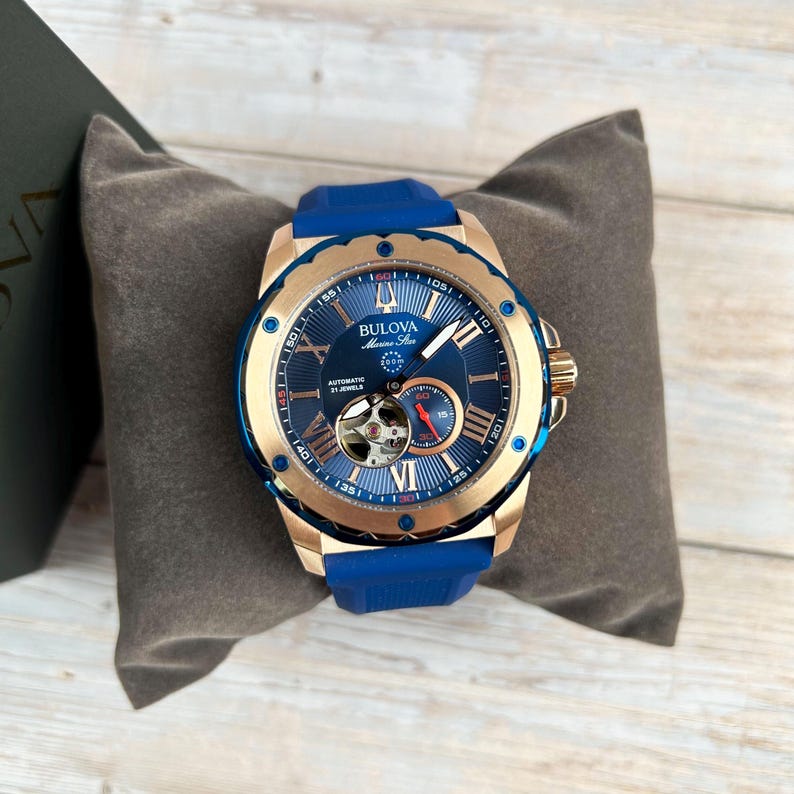 Pu&ograve; includere: Un orologio automatico Bulova Marine Star con quadrante blu, cassa color oro e cinturino in gomma blu. L'orologio presenta numeri romani e un movimento a vista. L'orologio &egrave; esposto su un cuscino grigio.