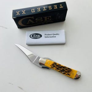 May include: A Case XX pocket knife with a yellow and brown handle, a silver blade, and a black box with the word "TESTED" on top. A white paper with the Case XX logo and "Product Quality Information" is also present.