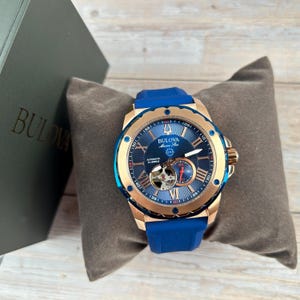 Pu&ograve; includere: Un orologio automatico Bulova Marine Star con cinturino in gomma blu. L'orologio ha una cassa color oro e un quadrante blu con numeri romani. L'orologio &egrave; esposto su un cuscino grigio accanto a una scatola grigia con il logo Bulova.