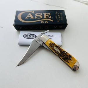 May include: A Case XX pocket knife with a yellow and brown handle, open to reveal a silver blade. The knife is displayed with its original packaging, including a black box and a white paper sleeve.