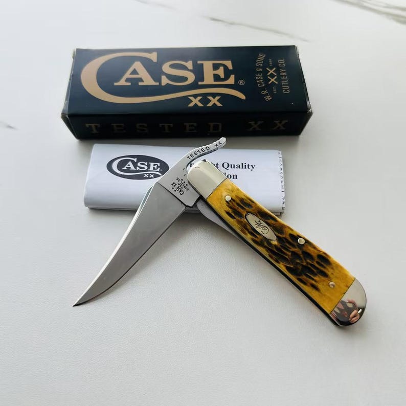 May include: A Case XX pocket knife with a yellow and brown handle, open to reveal a silver blade. The knife is displayed with its original packaging, including a black box and a white paper sleeve.