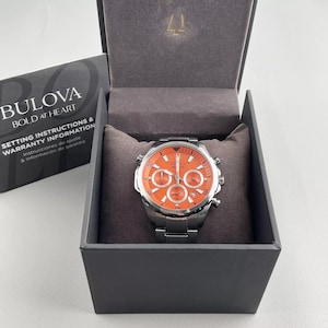 May include: A Bulova watch with an orange face and silver band is displayed in its original box. The watch face has multiple sub-dials and silver accents. The box is dark gray with a gray interior. The Bulova logo and text are visible on the box.