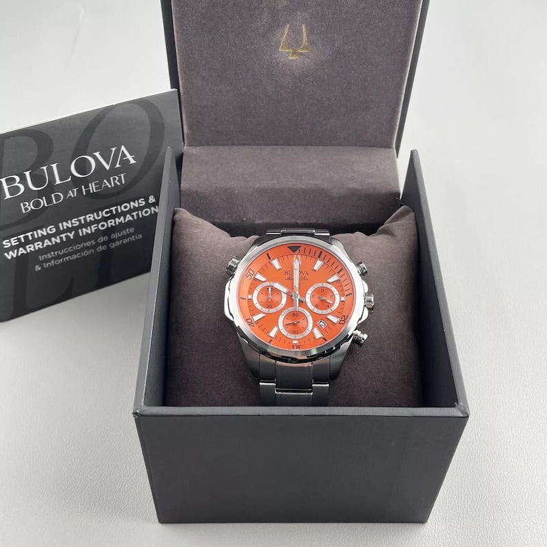 May include: A Bulova watch with an orange face and silver band is displayed in its original box. The watch face has multiple sub-dials and silver accents. The box is dark gray with a gray interior. The Bulova logo and text are visible on the box.
