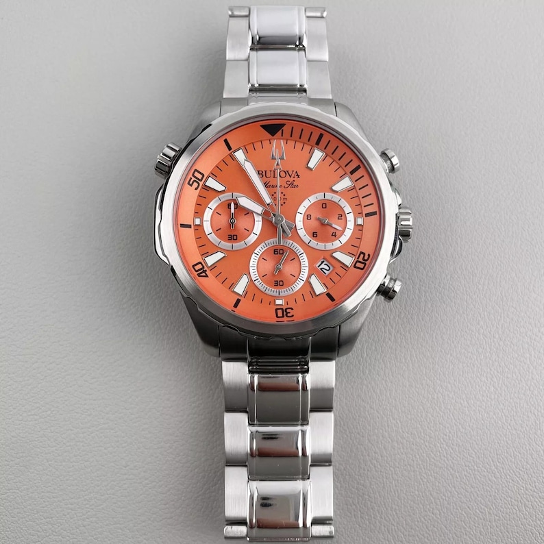 May include: A stainless steel Bulova watch with an orange face and a matching stainless steel band. The watch face features three sub-dials and the Bulova logo. The watch has a sporty design.