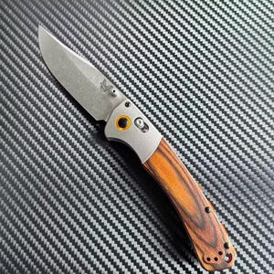 May include: A folding knife with a stainless steel blade and a wooden handle. The handle has a dark brown and reddish brown wood grain pattern. The blade has a small logo on it.