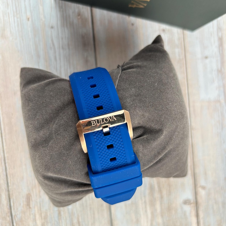 Pu&ograve; includere: Cinturino blu Bulova con fibbia color oro. Il cinturino &egrave; realizzato in materiale strutturato, simile alla gomma. La fibbia &egrave; stampata con il marchio Bulova. Il cinturino &egrave; appoggiato su un cuscino grigio.