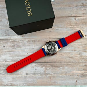 Pu&ograve; includere: Un orologio Bulova con cinturino rosso e blu, esposto su una superficie di legno. L'orologio ha un retro trasparente, che rivela il meccanismo interno. Il cinturino &egrave; rosso con dettagli blu. Una scatola verde scuro con la scritta "BULOV" &egrave; sullo sfondo.