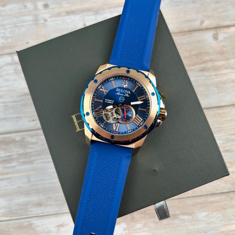 Pu&ograve; includere: Un orologio automatico Bulova Marine Star con quadrante blu e cassa color oro. L'orologio presenta numeri romani e un cinturino in gomma blu. L'orologio &egrave; presentato su una scatola grigio scuro.