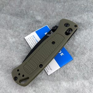 Peut inclure: Un couteau pliant vert olive avec un manche texturé et une lame noire. Le couteau a du matériel noir et un trou de lanière. Une étiquette bleue et blanche est partiellement visible.
