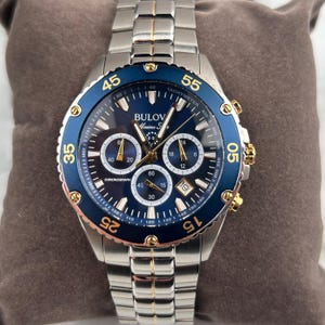 May include: A Bulova Marine Star chronograph watch with a stainless steel band and a blue bezel. The watch face is dark blue with gold accents and multiple sub-dials. The watch is displayed on a brown cushion.