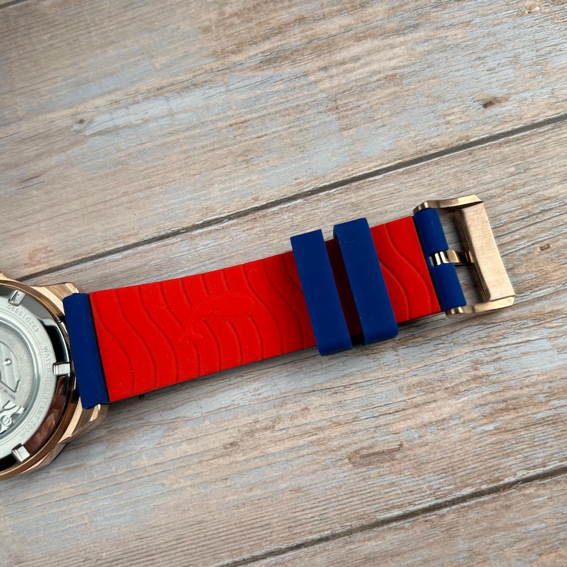 Pu&ograve; includere: Primo piano di un orologio da polso con cinturino in silicone rosso e blu. Il cinturino ha una trama a onde ed &egrave; fissato con una fibbia color oro. Il quadrante dell'orologio &egrave; parzialmente visibile, mostrando un quadrante argentato.