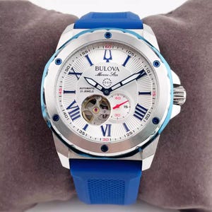 May include: A Bulova Marine Star automatic watch with a silver face, blue accents, and a blue silicone strap. The watch features Roman numeral hour markers, a visible movement, and a small seconds dial. The watch is water-resistant to 200m.