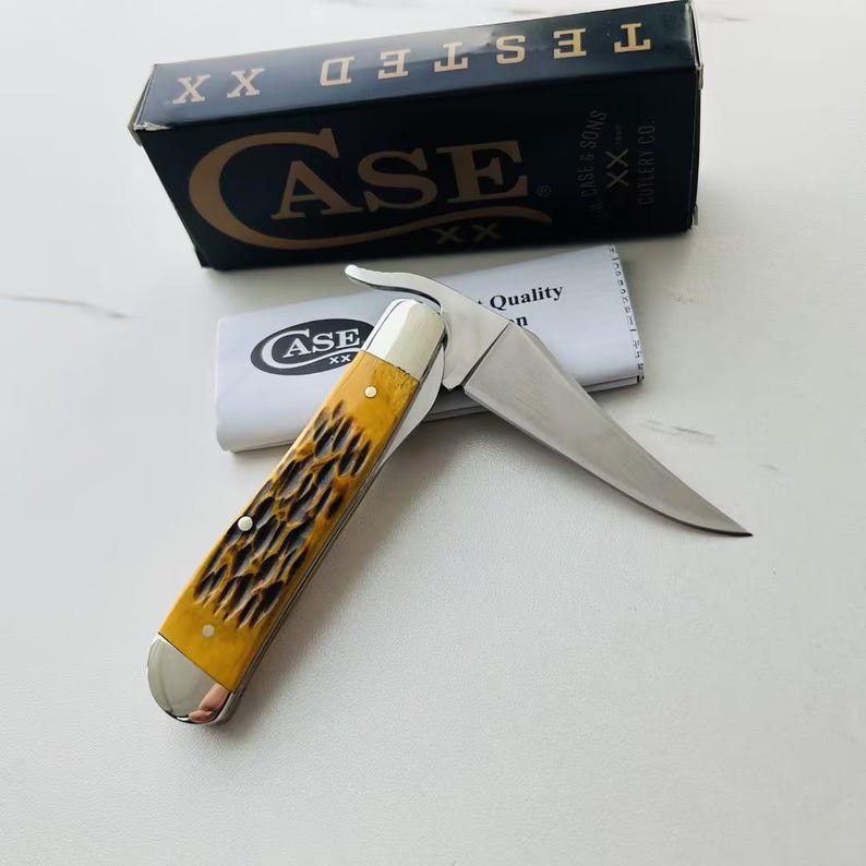 May include: A Case XX folding knife with a yellow and brown handle and a silver blade. The knife is partially open, revealing the sharp edge. A black box with gold lettering that reads "CASE" and "TESTED XX" is in the background.