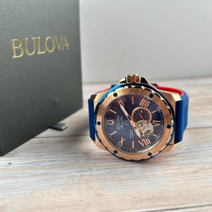 Pu&ograve; includere: Un orologio automatico Bulova Marine Star con quadrante blu, cassa color oro e cinturino in gomma blu. L'orologio presenta numeri romani e un movimento a vista. L'orologio &egrave; esposto accanto a una scatola verde scuro con il logo Bulova.