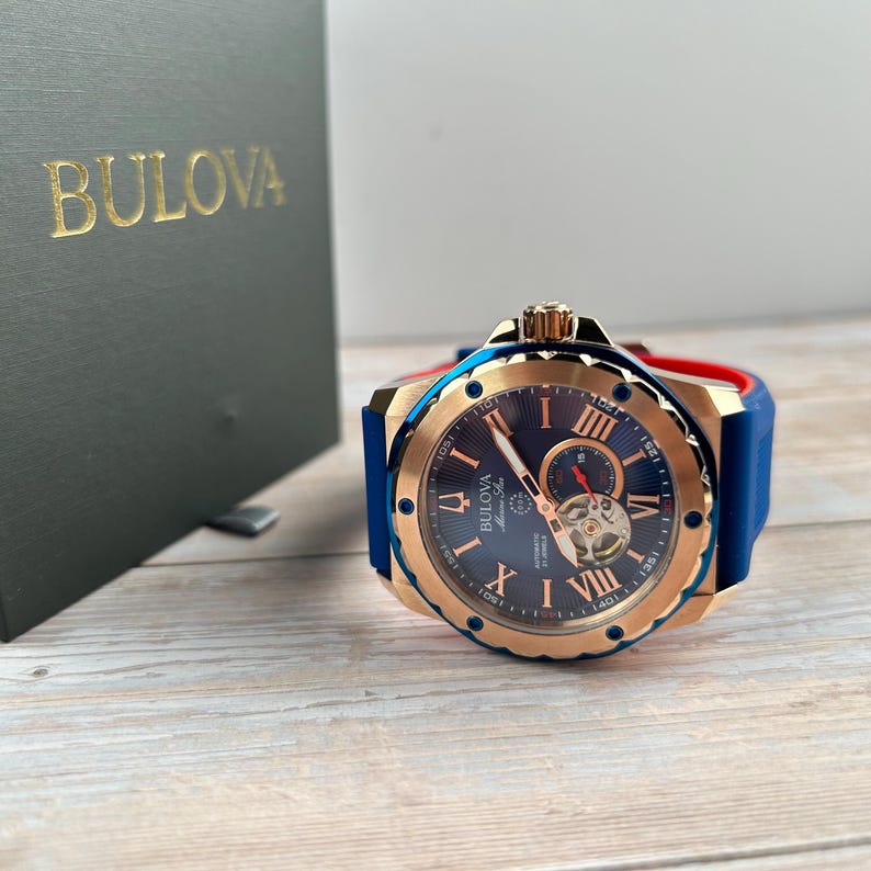 Pu&ograve; includere: Un orologio automatico Bulova Marine Star con quadrante blu, cassa color oro e cinturino in gomma blu. L'orologio presenta numeri romani e un movimento a vista. L'orologio &egrave; esposto accanto a una scatola verde scuro con il logo Bulova.