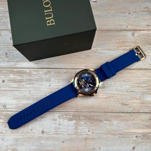 Pu&ograve; includere: Un orologio Bulova blu con lunetta color oro e cinturino in silicone blu. Il quadrante dell'orologio &egrave; blu con dettagli dorati e meccanismi interni visibili. L'orologio &egrave; esposto con la sua scatola verde su una superficie di legno.