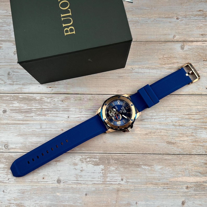 Pu&ograve; includere: Un orologio Bulova blu con lunetta color oro e cinturino in silicone blu. Il quadrante dell'orologio &egrave; blu con dettagli dorati e meccanismi interni visibili. L'orologio &egrave; esposto con la sua scatola verde su una superficie di legno.