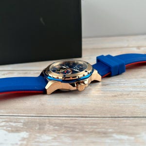 Pu&ograve; includere: Un orologio da polso di lusso con quadrante blu, cassa color oro e cinturino in silicone blu e rosso. L'orologio presenta un design complesso del quadrante ed &egrave; appoggiato su una superficie di legno.
