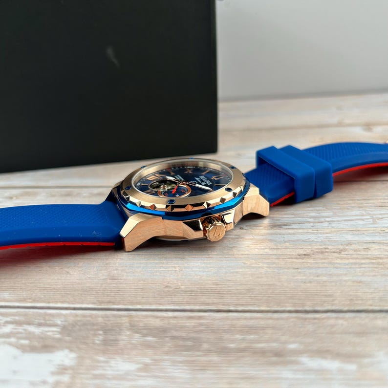 Pu&ograve; includere: Un orologio da polso di lusso con quadrante blu, cassa color oro e cinturino in silicone blu e rosso. L'orologio presenta un design complesso del quadrante ed &egrave; appoggiato su una superficie di legno.