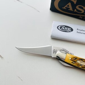 May include: A Case XX pocket knife with a polished steel blade and a yellow handle. The knife is partially open, displaying the blade. The handle has a textured appearance. A product information sheet and box are in the background.