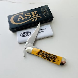 May include: A Case XX folding knife with a yellow and brown handle, a silver blade, and a curved metal piece. The knife is next to a black box with the Case XX logo and the words "TESTED" and "CUTLERY CO."