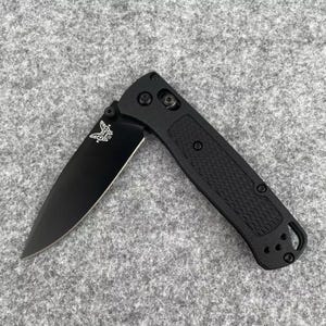 May include: Black folding knife with a partially open blade. The blade has a silver butterfly logo. The handle is black with a textured grip. The knife is on a gray surface.