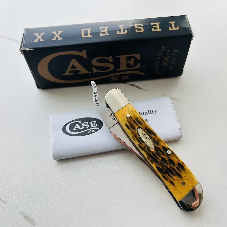 May include: A Case pocket knife with a yellow and brown handle, a silver blade, and a silver bolster. The knife is partially open, with a white paper and a black box in the background. The box has the word "CASE" in gold lettering.
