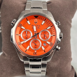 May include: A stainless steel Bulova watch with an orange face. The watch features three sub-dials, a date window, and luminous hands. The watch has a silver band and a black triangle at the top of the face.