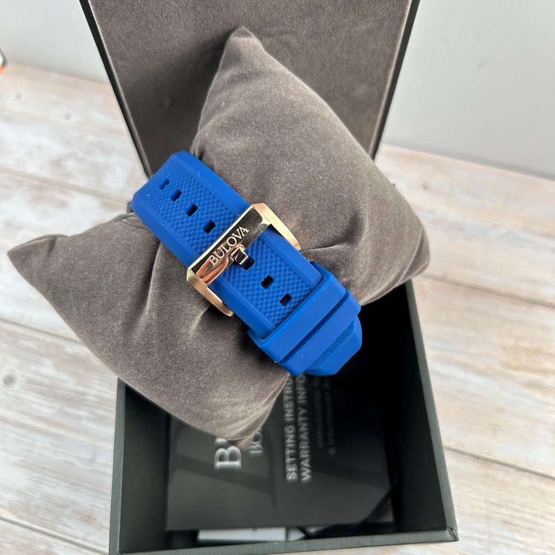 Pu&ograve; includere: Un cinturino blu Bulova con fibbia color oro, appoggiato su un cuscino grigio all'interno di una scatola nera. La fibbia &egrave; incisa con il marchio Bulova. Il cinturino &egrave; realizzato in gomma strutturata.